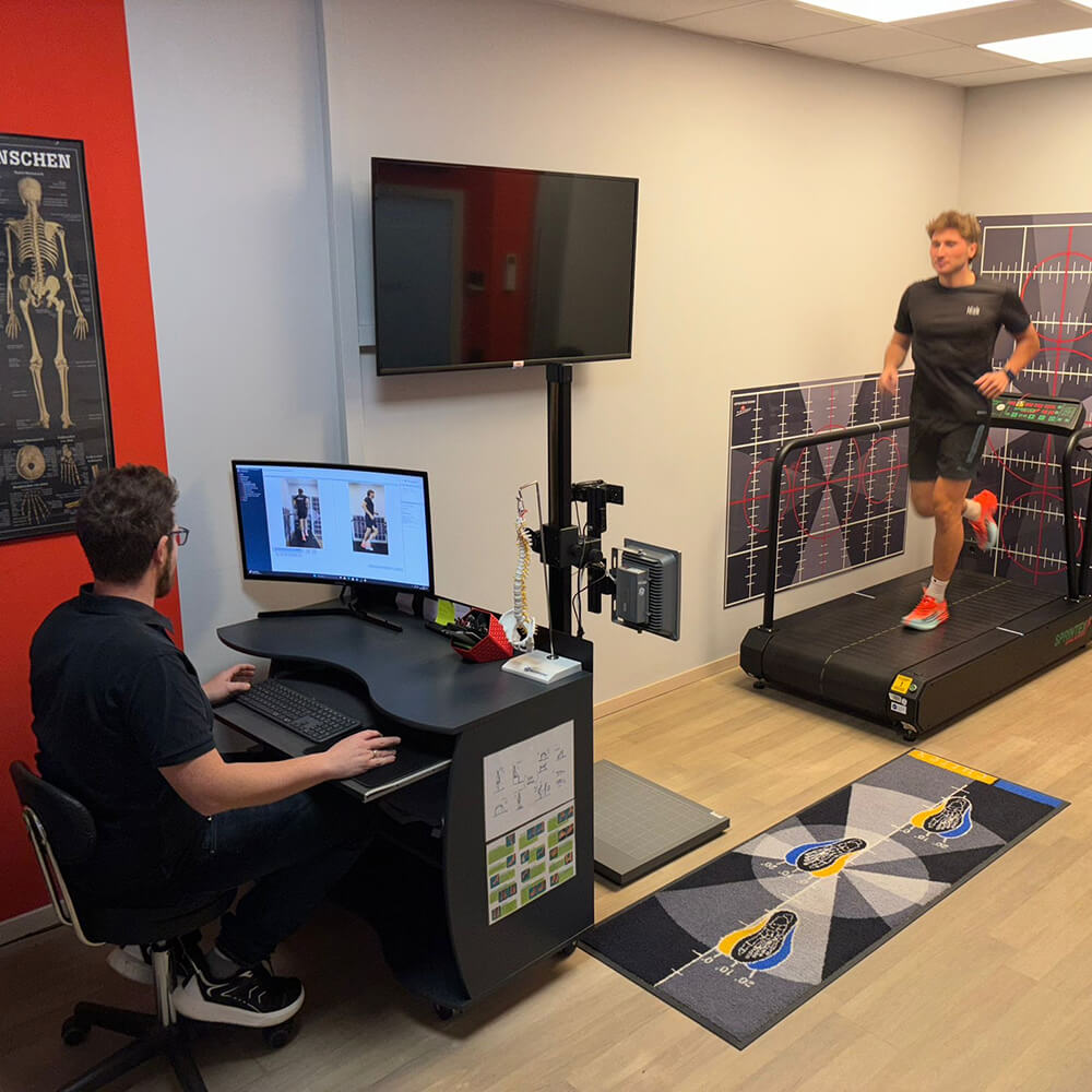 Übersicht eines Analyseraums mit Laufband, Monitoren und Fachpersonal, das die Laufbewegung eines Sportlers auswertet